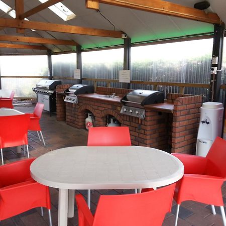 Taupo Debretts Spa Resort Экстерьер фото The photo shows an outdoor kitchen area with a covered space. There are several barbecue grills set up on a brick counter. In the foreground, there are round tables with white tops and bright red chairs arranged around them. A Coca-Cola vending machi