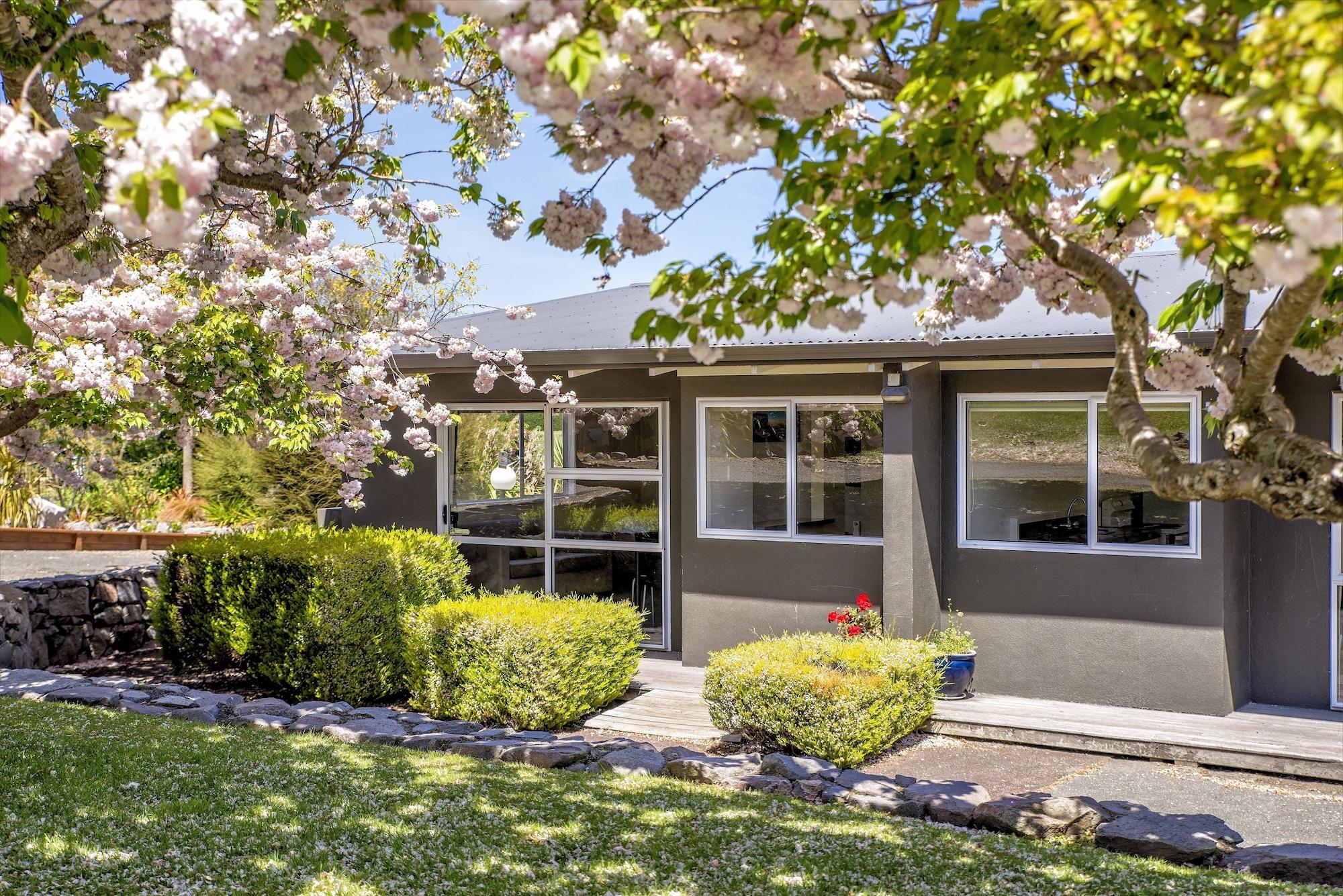 Taupo Debretts Spa Resort Экстерьер фото The photo shows a charming house surrounded by vibrant greenery and blooming trees. There are pink flowers from the trees adding a pop of color to the scene. The house features large windows that allow natural light to fill the interior. A well-maint