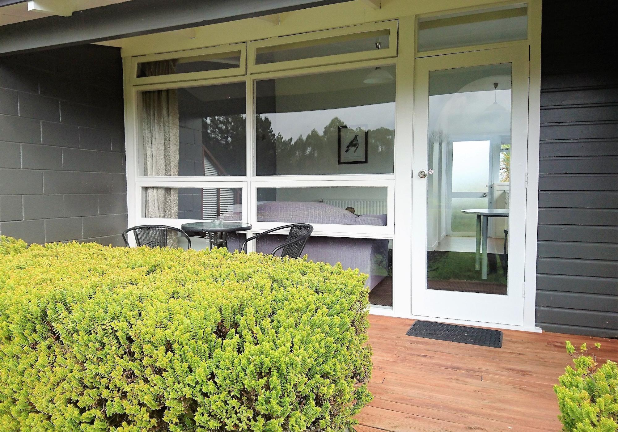Taupo Debretts Spa Resort Экстерьер фото The photo shows a patio area in front of a building. There are green shrubs or bushes lining the foreground, creating a natural border. In the center, there is a wooden deck with a small table and two chairs, suggesting a cozy seating area. The door 