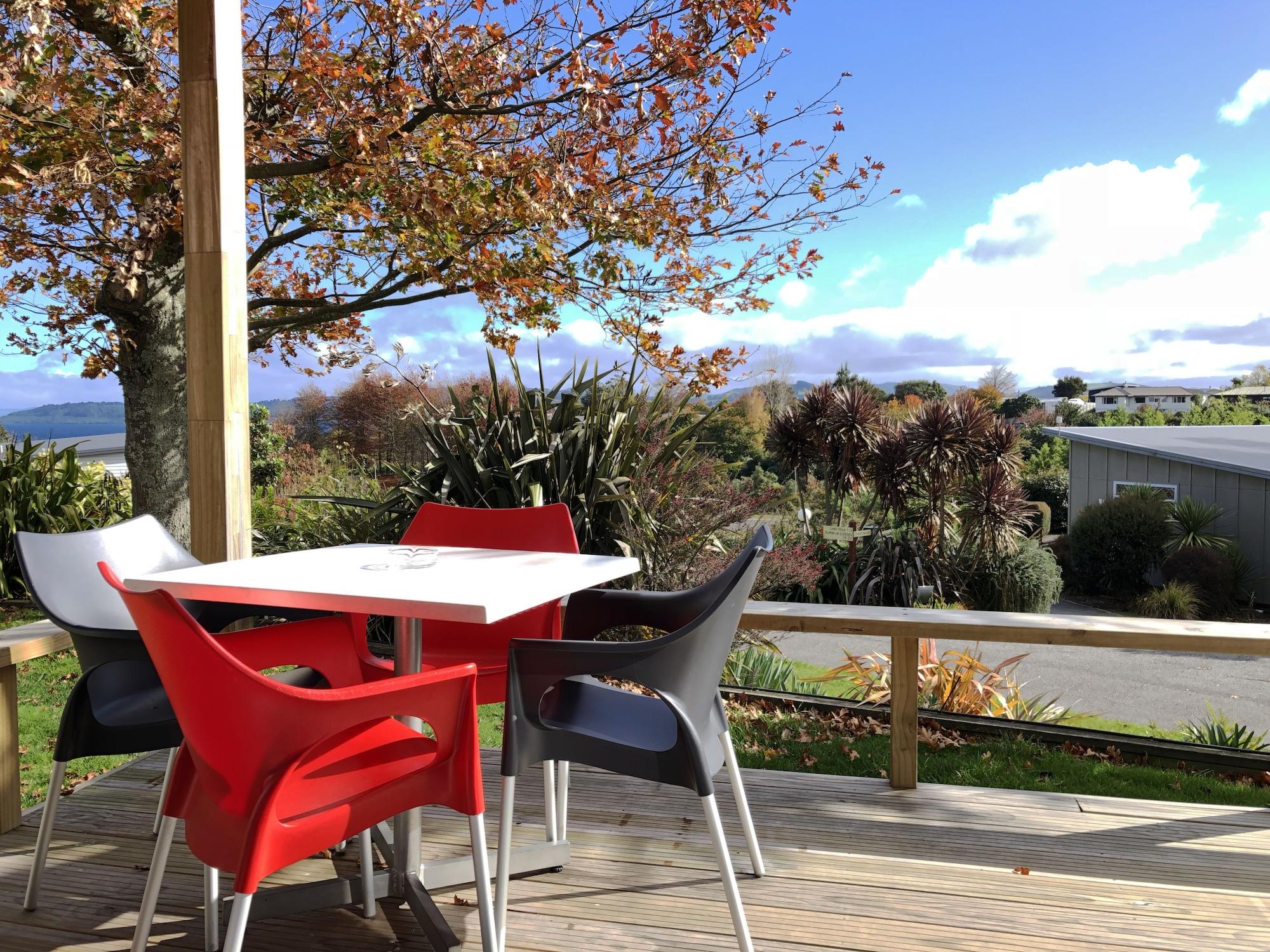 Taupo Debretts Spa Resort Экстерьер фото The photo shows a picturesque outdoor seating area on a deck. There is a table surrounded by several chairs, with a couple of them in red and the others in gray and white. In the background, there are trees displaying autumn foliage, and beyond that,