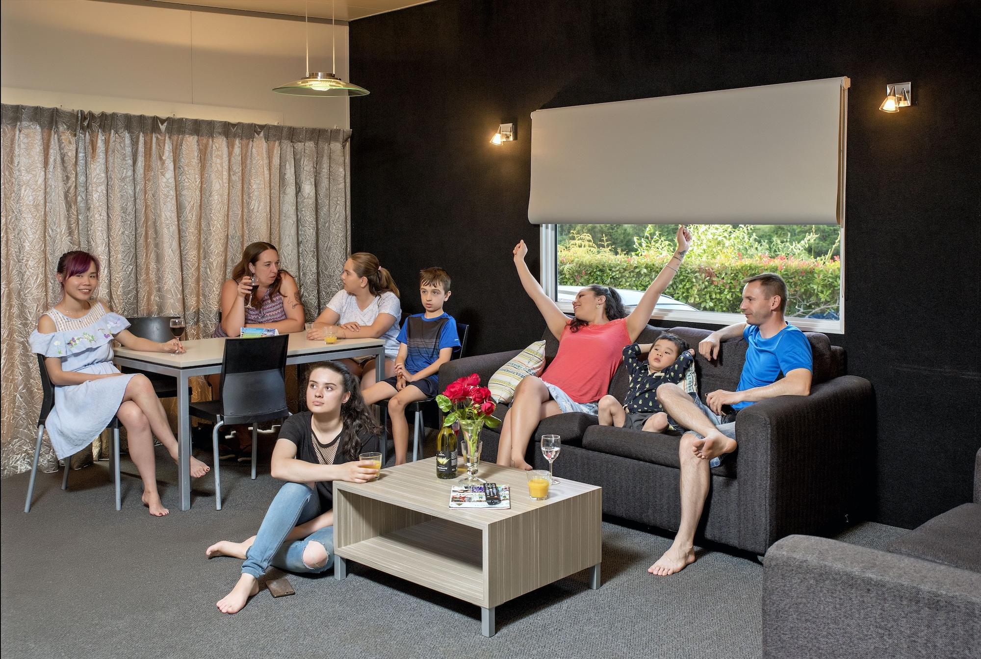 Taupo Debretts Spa Resort Экстерьер фото The photo shows a group of people gathered in a cozy living room. There are several adults and children interacting with each other. One woman in a red top appears to be celebrating or expressing excitement, while others are engaged in conversation o