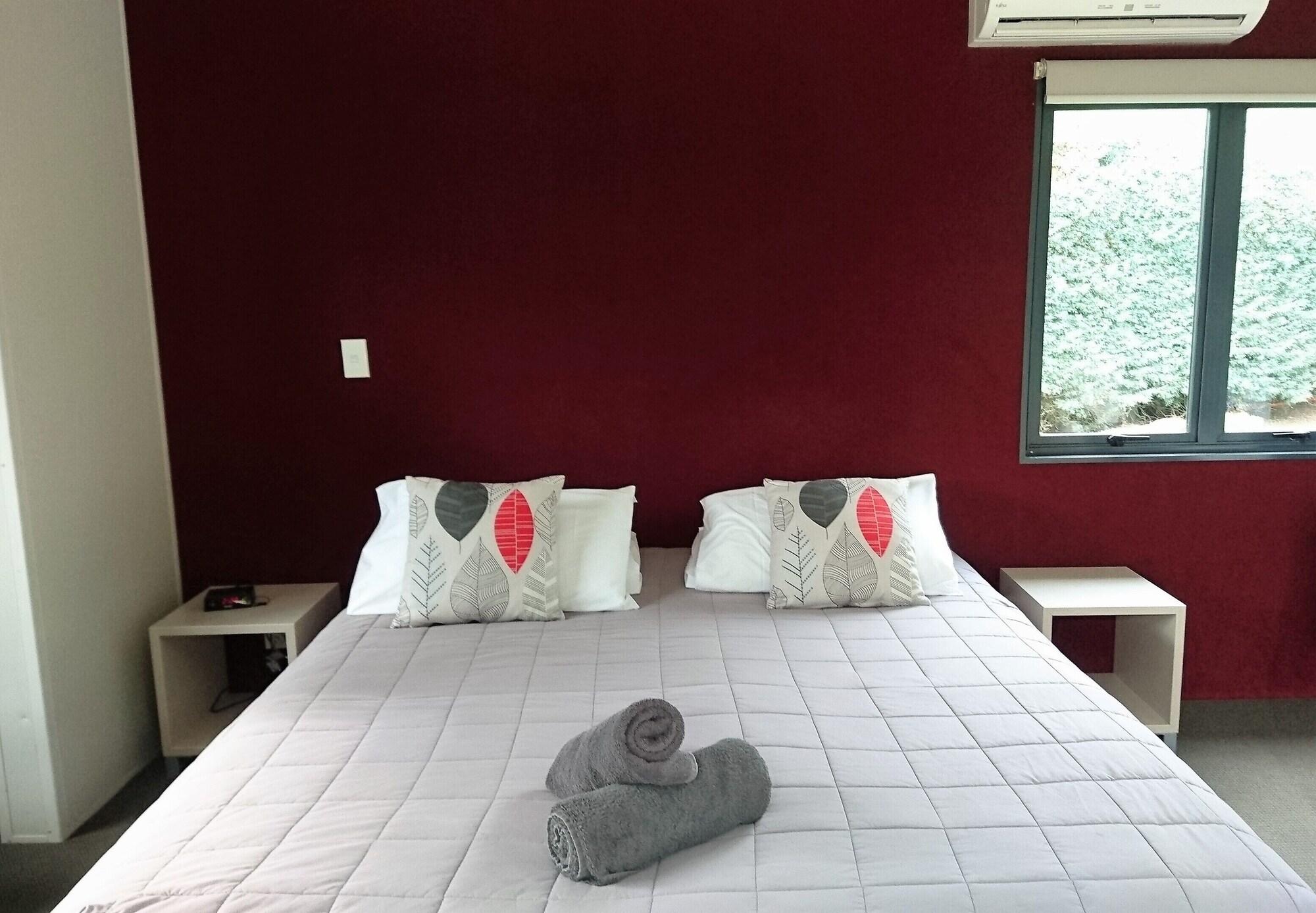 Taupo Debretts Spa Resort Экстерьер фото The photo shows a cozy bedroom featuring a large bed with a light-colored quilt. The bed has two decorative pillows with a simple design on each side. There are two bedside tables, one on each side of the bed. On the bed, there are rolled towels plac