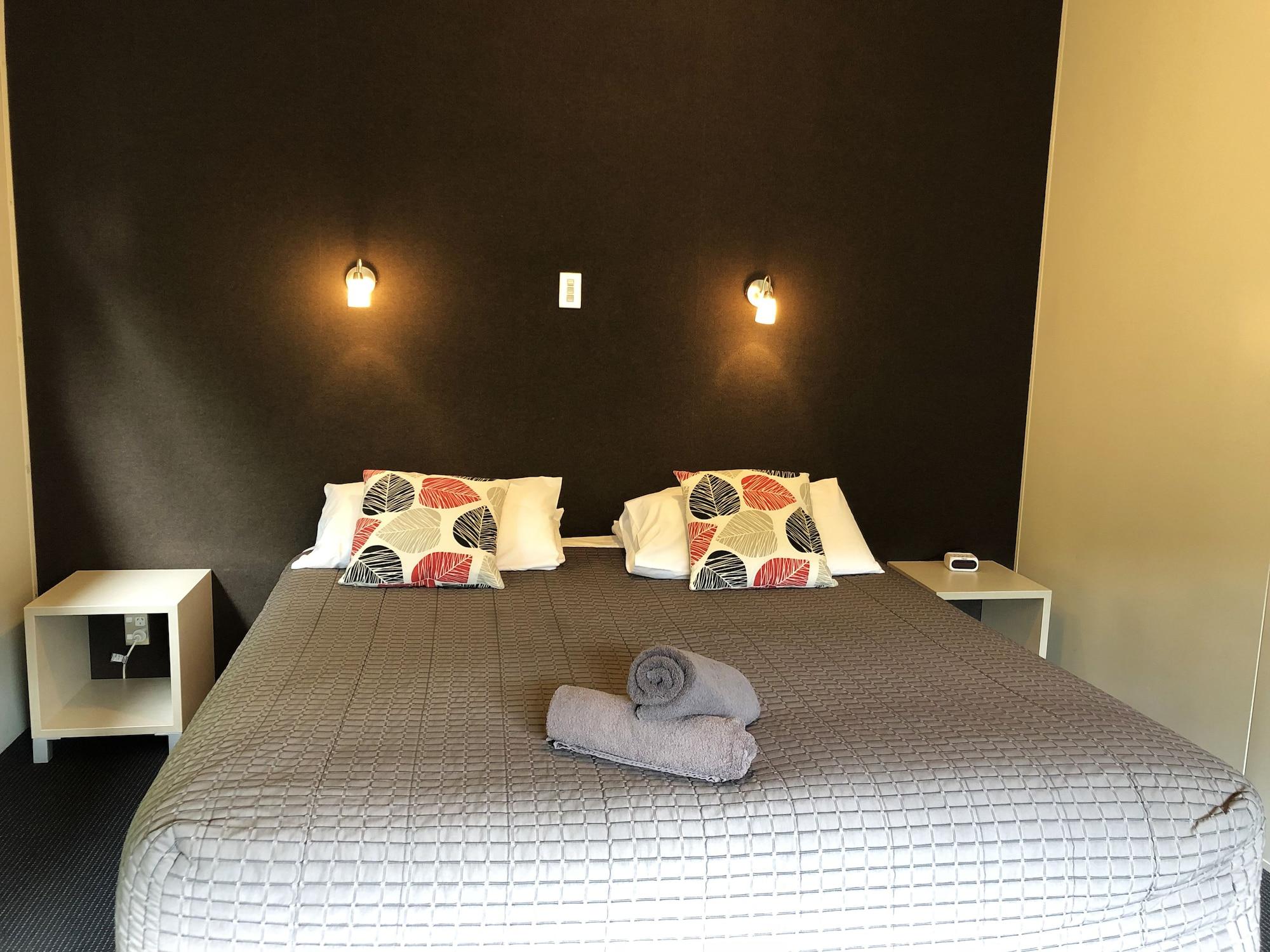 Taupo Debretts Spa Resort Экстерьер фото The photo shows a bedroom with a large bed that has a gray quilted cover and decorative pillows. The walls behind the bed are painted a dark color, and there are two small wall-mounted lamps providing light. On either side of the bed, there are small