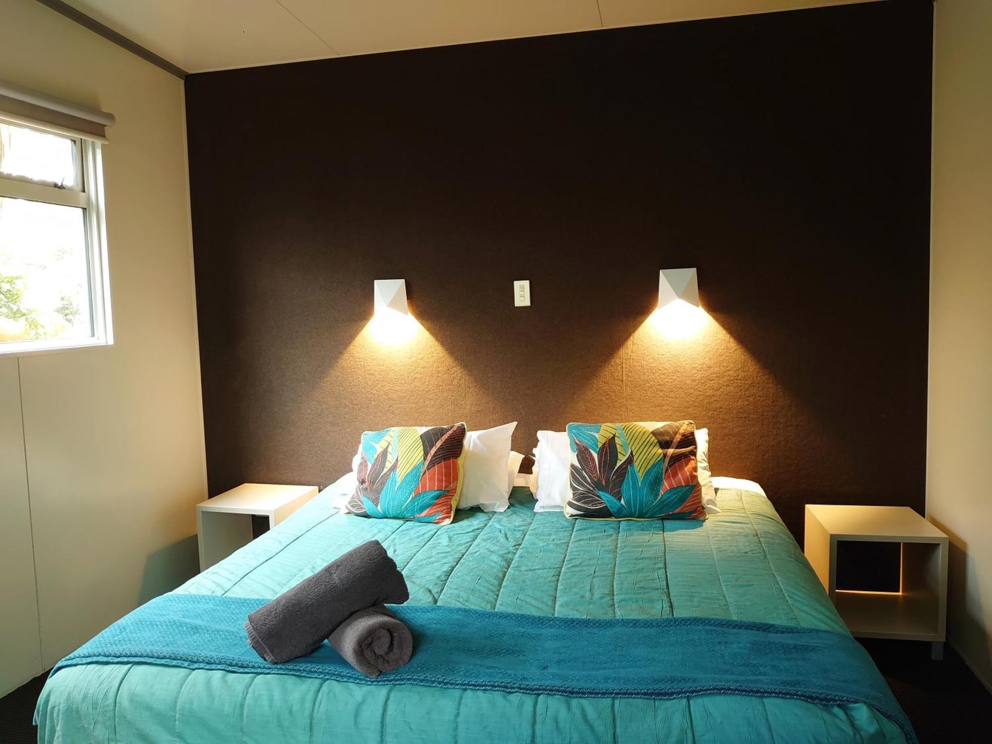 Taupo Debretts Spa Resort Экстерьер фото The image shows a bedroom with a modern and inviting layout. The focal point is a bed with a vibrant turquoise comforter, decorated with two colorful pillows featuring floral patterns. On either side of the bed, there are small nightstands with wall-