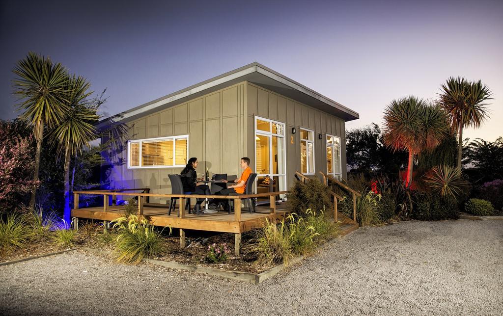 Taupo Debretts Spa Resort Экстерьер фото The photo depicts a modern, single-story house with a light-colored exterior. It has large windows and a sloped roof. In front of the house, there is a wooden deck where two people are seated, engaged in conversation or perhaps enjoying a meal. The a
