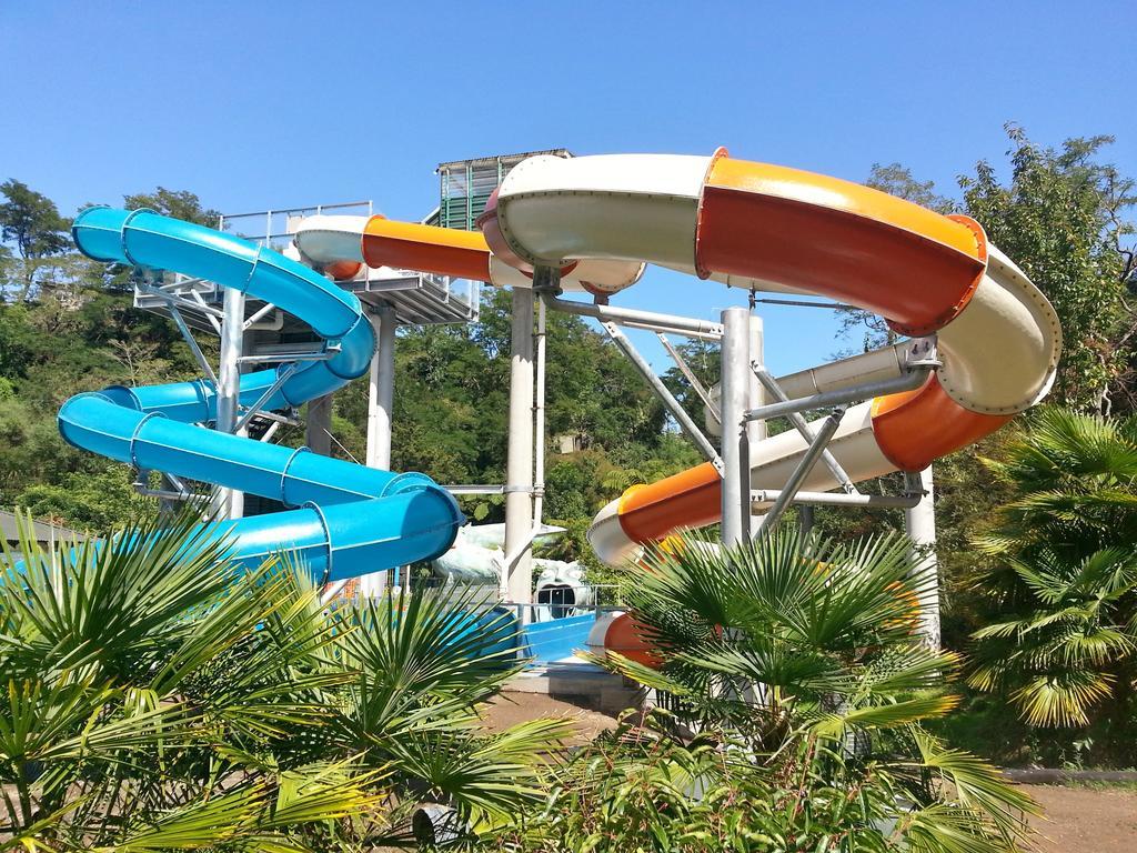 Taupo Debretts Spa Resort Экстерьер фото The photo shows a water park featuring two large water slides. One slide is blue and curves gracefully, while the other is orange and white, twisting as it descends. They are supported by a metal structure and surrounded by lush greenery, including p