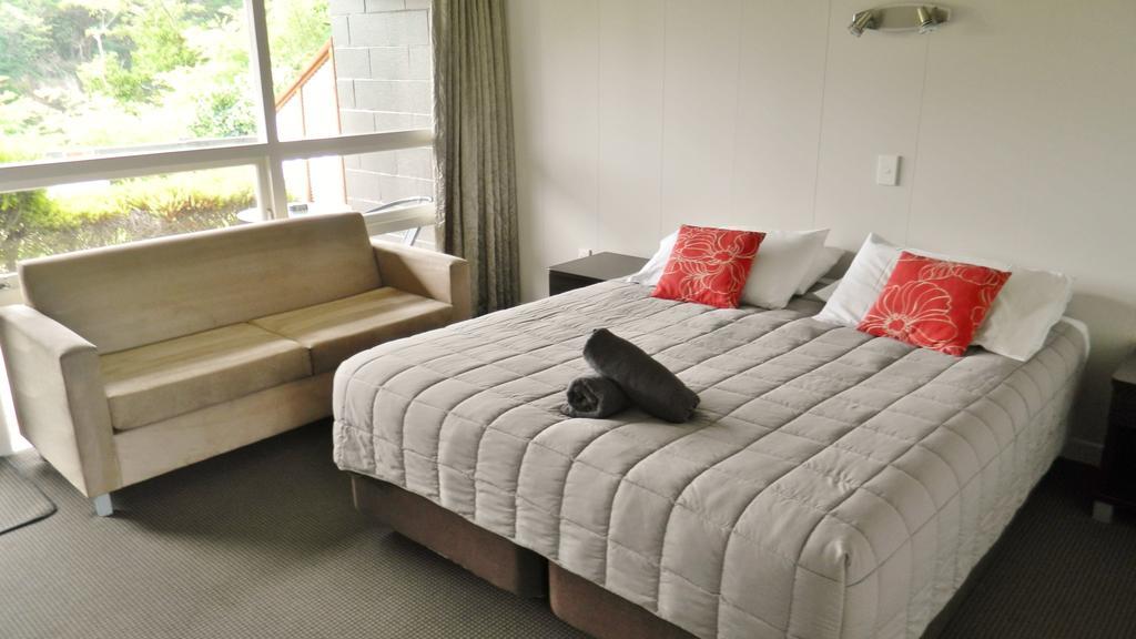 Taupo Debretts Spa Resort Экстерьер фото The photo shows a cozy hotel or guest room. There is a large bed with gray bedding and two red decorative pillows. At the foot of the bed, there is a rolled-up towel. Next to the bed, there's a small bedside table and a light fixture on the wall. To 