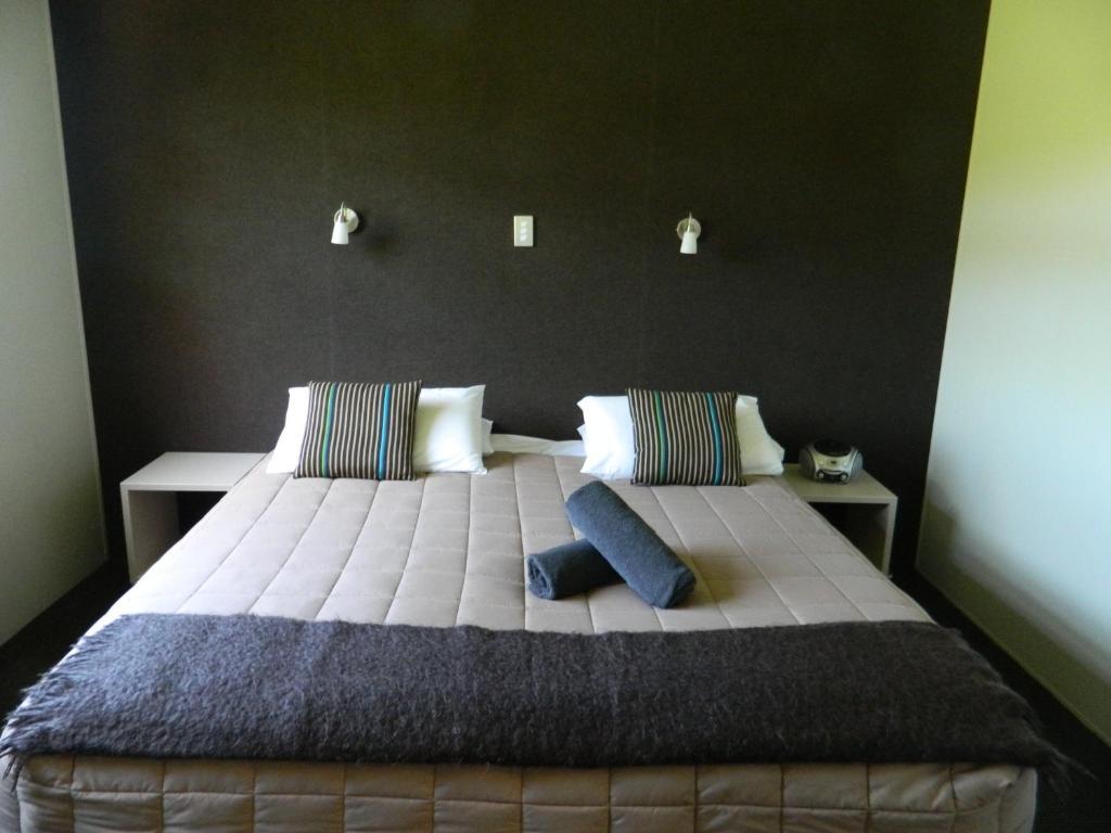 Taupo Debretts Spa Resort Экстерьер фото The photo shows a modern bedroom featuring a large bed with a neutral-colored quilt and decorative pillows. There are two bedside tables on either side of the bed. The wall behind the bed is dark, creating a contrast with the lighter bedding. Additio
