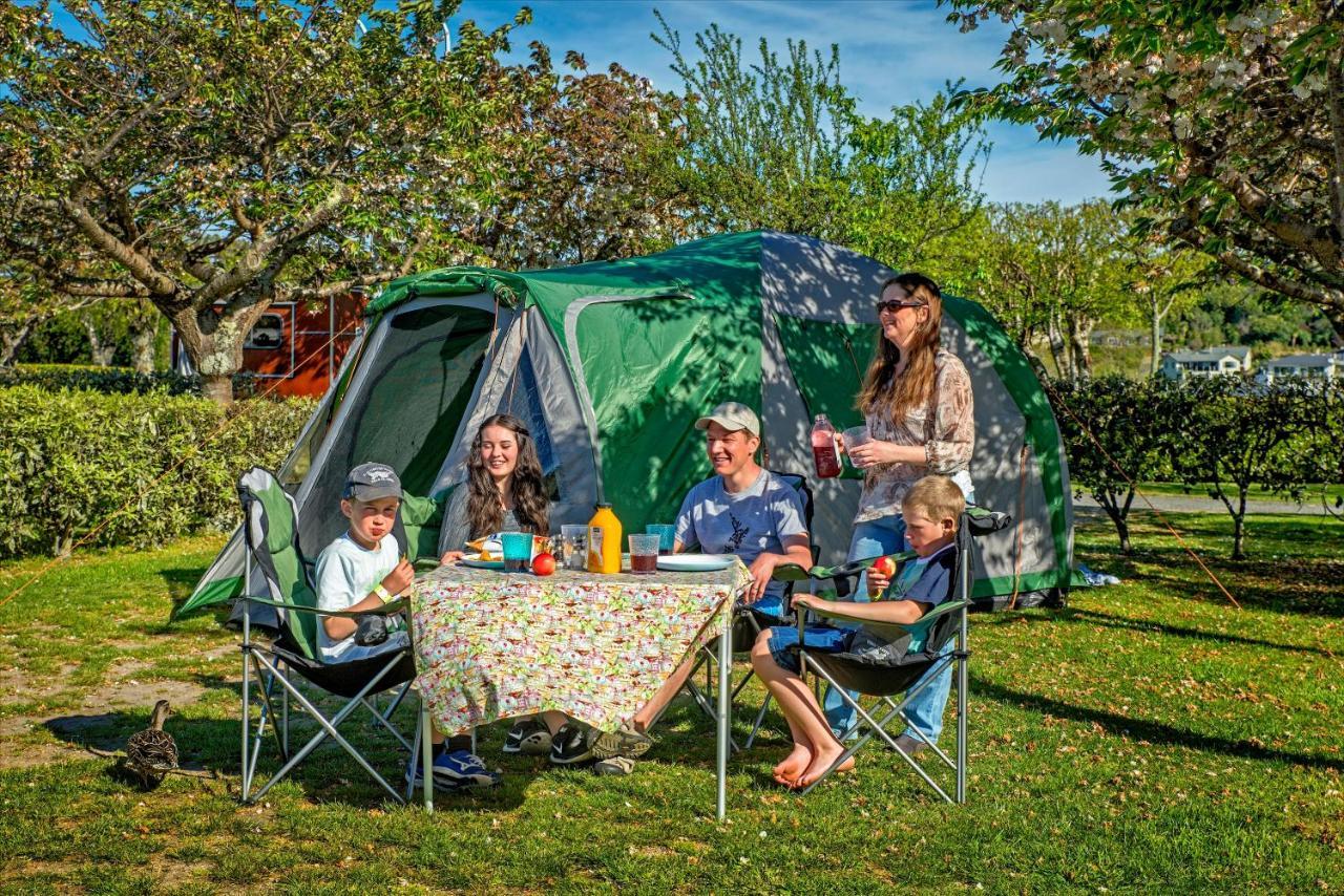 Taupo Debretts Spa Resort Экстерьер фото The photo shows a group of five people enjoying a picnic outside a green tent in a park-like setting. They are seated around a table covered with a floral tablecloth, which has various food and drink items on it. The group includes two adults and thr