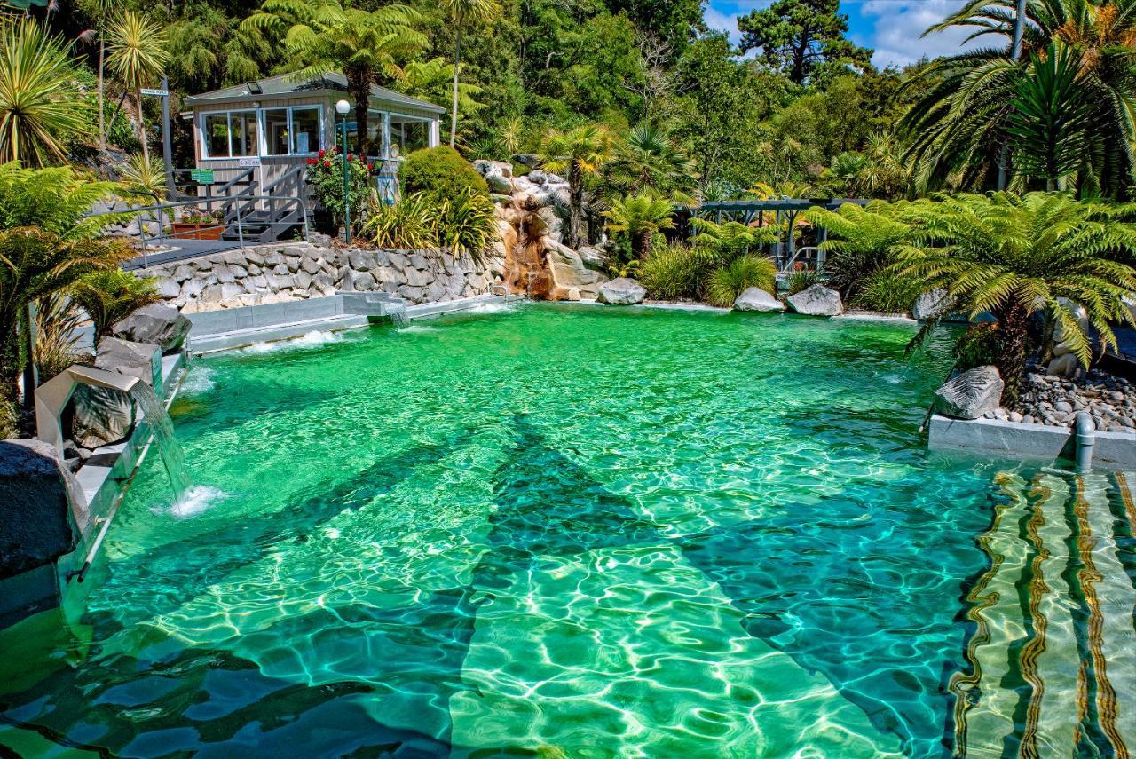 Taupo Debretts Spa Resort Экстерьер фото The photo showcases a vibrant, natural-looking pool with crystal-clear, turquoise water. Surrounding the pool are lush green plants and palm trees, creating a tropical atmosphere. In the background, there is a white building, possibly a facility or a