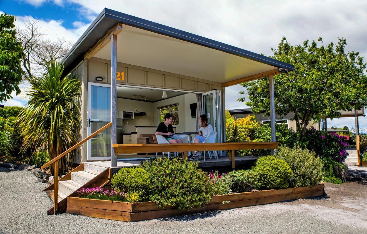 Taupo Debretts Spa Resort Экстерьер фото The photo depicts a modern small house or cabin, featuring a covered porch with a wooden railing. Two people are seated at a table on the porch, engaging in conversation. The exterior of the house is designed with large windows, allowing ample natura
